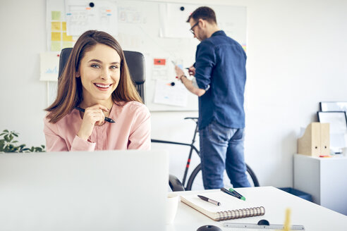 Porträt einer lächelnden Frau, die an einem Schreibtisch im Büro arbeitet, mit einem Kollegen im Hintergrund - BSZF00996