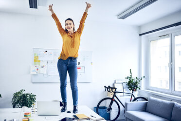 Happy young businesswoman cheering in office standing on desk - BSZF01029