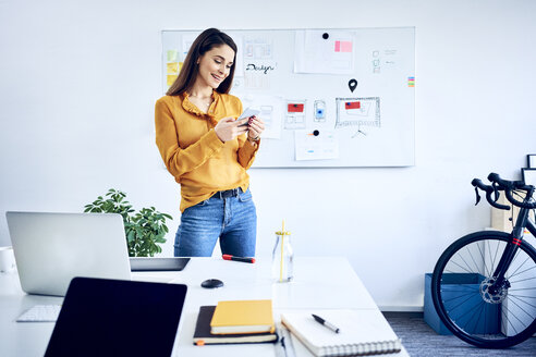 Lächelnde Geschäftsfrau hält Smartphone an Whiteboard im Büro - BSZF01038