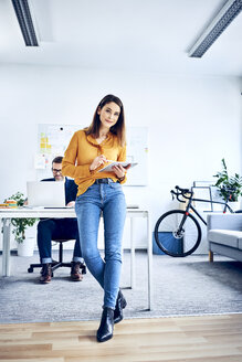 Porträt einer Geschäftsfrau, die im Büro ein Tablet benutzt, mit einem Kollegen im Hintergrund - BSZF01043