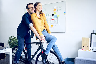 Happy colleagues riding bicycle together in office - BSZF01058