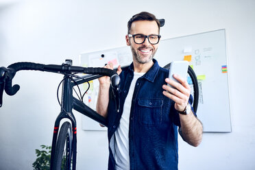 Lächelnder Geschäftsmann mit Fahrrad im Büro, der auf sein Smartphone schaut - BSZF01061