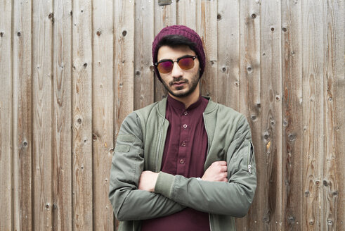 Portrait of serious bearded man in purple knit hat and shirt with arms crossed wearing sunglasses over wooden background - IGGF00892