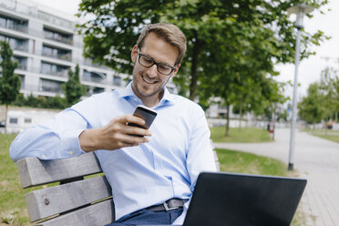 Junger Geschäftsmann sitzt auf einer Parkbank und benutzt Smartphone und Laptop - KNSF05572