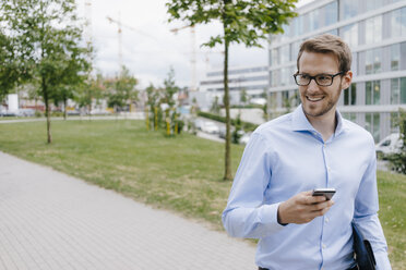 Young businessman walking in park, using smartphone - KNSF05573