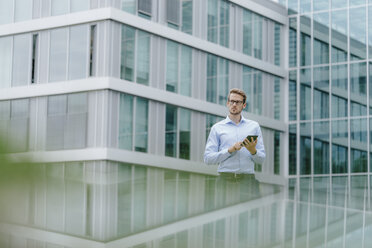 Young businessman standing in front of modern office building, usind digital tablet - KNSF05581
