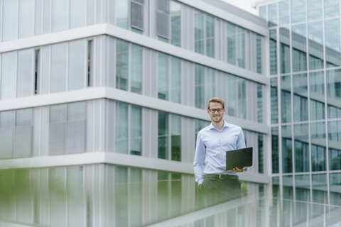 Junger Geschäftsmann steht vor einem modernen Bürogebäude und benutzt einen Laptop, lizenzfreies Stockfoto