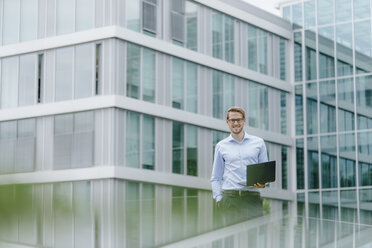 Junger Geschäftsmann steht vor einem modernen Bürogebäude und benutzt einen Laptop - KNSF05582