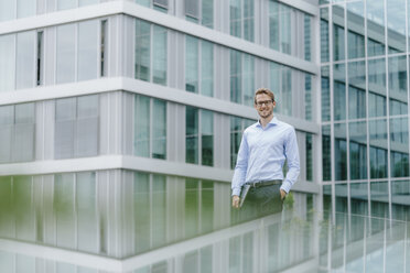 Young businessman standing in front of modern office building, carrying laptop - KNSF05583