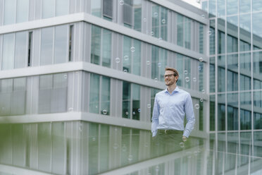 Young businessman standing in front of modern office building, watching soap bubbles - KNSF05584