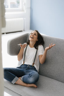 Hübsche Frau sitzt auf der Couch, benutzt ihr Smartphone und lacht - KNSF05616