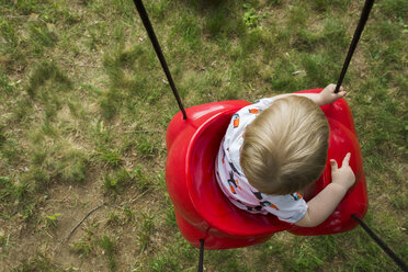 Draufsicht auf einen kleinen Jungen, der auf einer roten Plastikschaukel auf einem Spielplatz schaukelt - CAVF60638