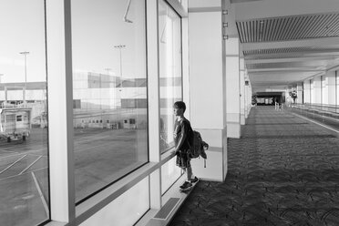 Side view of boy with backpack looking through window while standing at airport - CAVF60643