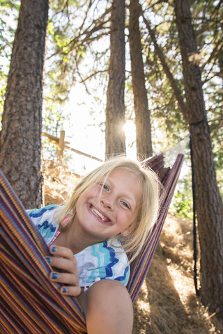 Porträt eines glücklichen Mädchens, das in einer Hängematte gegen Bäume im Wald sitzt, lizenzfreies Stockfoto