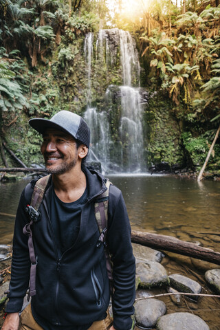 Lächelnder Wanderer, der vor einem Wasserfall im Wald steht, lizenzfreies Stockfoto