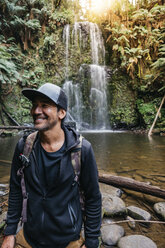 Lächelnder Wanderer, der vor einem Wasserfall im Wald steht - CAVF60668