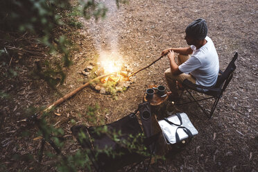 High angle view of hiker sitting on camping chair by campfire in forest - CAVF60670