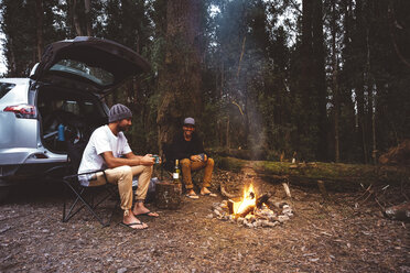 Freunde, die auf Campingstühlen am Lagerfeuer im Wald sitzen und etwas trinken - CAVF60671