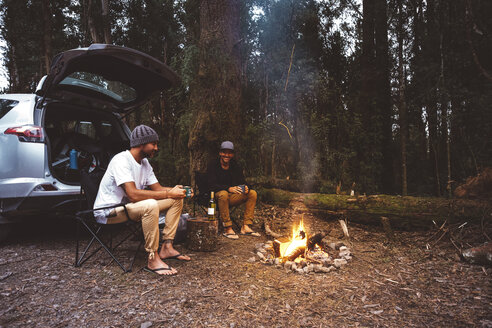 Freunde, die auf Campingstühlen am Lagerfeuer im Wald sitzen und etwas trinken - CAVF60671