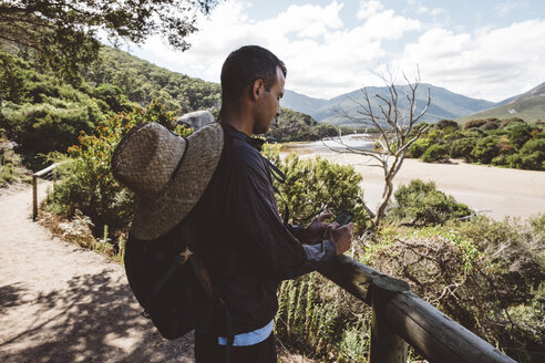 Seitenansicht eines Wanderers mit Rucksack und Strohhut, der ein Mobiltelefon benutzt, während er im Wald im Wilsons Promontory National Park steht - CAVF60679