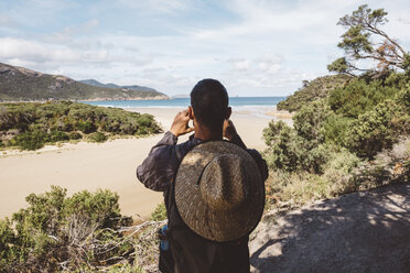 Rear view of hiker with backpack and straw hat photographing sea while standing in forest at Wilsons Promontory National Park - CAVF60680