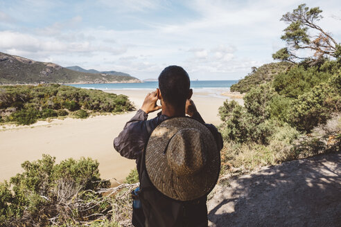 Rückansicht eines Wanderers mit Rucksack und Strohhut, der das Meer fotografiert, während er im Wald im Wilsons Promontory National Park steht - CAVF60680
