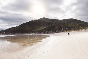 Mid distance view of hiker walking at beach towards mountains against cloudy sky - CAVF60686