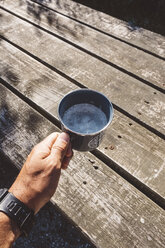 Abgeschnittene Hand eines Wanderers, der ein schaumiges Getränk in einem Becher über einem Holztisch hält - CAVF60695