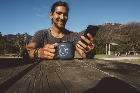 Lächelnder Wanderer, der eine Tasse hält, während er sein Handy auf einem Holztisch im Wilsons Promontory National Park benutzt - CAVF60696