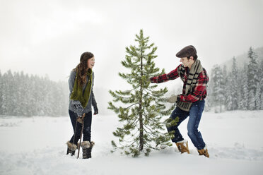 Happy couple standing by pine tree on snow covered field against sky in forest - CAVF60732