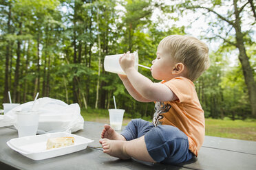 Full length of cute baby boy drinking drink while sitting on picnic tablet at park - CAVF60790