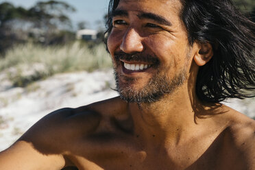 Nahaufnahme eines glücklichen Mannes ohne Hemd, der wegschaut, während er an einem sonnigen Tag am Hyams Beach sitzt - CAVF60799