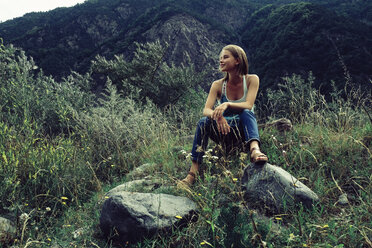Low angle view of woman looking away while sitting on grassy field against mountain - CAVF60806
