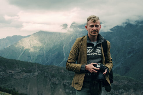 Porträt eines selbstbewussten Mannes, der eine Kamera hält, während er auf einem Berg vor einem bewölkten Himmel steht - CAVF60810