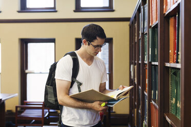 Selbstbewusster Mann mit Rucksack, der ein Buch liest, während er an einem Regal in einer Bibliothek steht - CAVF60814