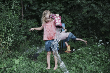 Schwester gießt in voller Länge Wasser aus Gummistiefeln, während Bruder inmitten von Pflanzen im Park steht - CAVF60847