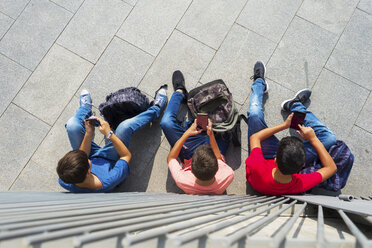 Hochformatige Ansicht von Freunden, die Smartphones benutzen, während sie an einem sonnigen Tag auf einem Gehweg an einer Mauer sitzen - CAVF60874