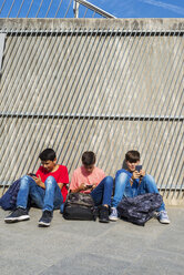 Freunde benutzen Mobiltelefone, während sie an einem sonnigen Tag auf dem Gehweg an der Wand sitzen - CAVF60875