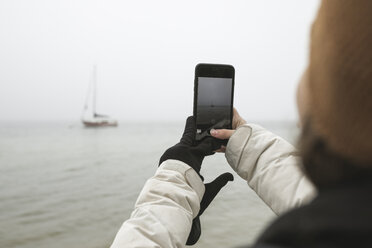 Junge Frau, die mit ihrem Smartphone im Winter ein Schiff auf dem Meer fotografiert - CAVF60879