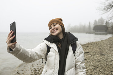 Lächelnde junge Frau nimmt Selfie mit Smartphone am Strand im Winter - CAVF60880