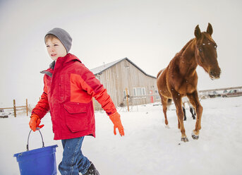 Junge mit Eimer läuft auf Schnee gegen Pferd auf Bauernhof im Winter - CAVF60887
