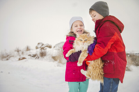 Glückliche Geschwister, die eine Katze tragen, während sie im Winter im Schnee stehen, lizenzfreies Stockfoto