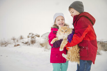 Glückliche Geschwister, die eine Katze tragen, während sie im Winter im Schnee stehen - CAVF60888