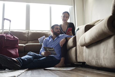 Junges Paar, das ein Mobiltelefon benutzt, während es im Wohnzimmer eines Miethauses sitzt - CAVF60901