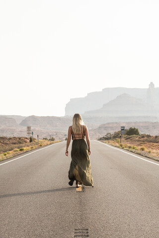 Rückansicht einer Frau, die auf der Straße gegen den klaren Himmel im Monument Valley Tribal Park läuft, an einem sonnigen Tag, lizenzfreies Stockfoto