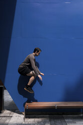 Seitenansicht eines Mannes, der auf einer Holzbank gegen eine blaue Wand springt, während eines sonnigen Tages - CAVF60961