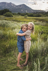 Portrait of happy sisters embracing while standing on grassy field in forest during sunset - CAVF61005