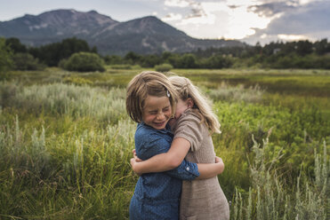 Happy sisters embracing while standing on grassy field in forest during sunset - CAVF61007