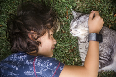 Hoher Blickwinkel des Jungen, der eine Katze streichelt, während er auf einer Wiese im Hof liegt - CAVF61026