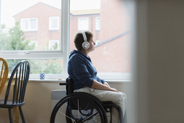 Nachdenkliche junge Frau im Rollstuhl, die mit Kopfhörern am Fenster Musik hört - CAIF22615